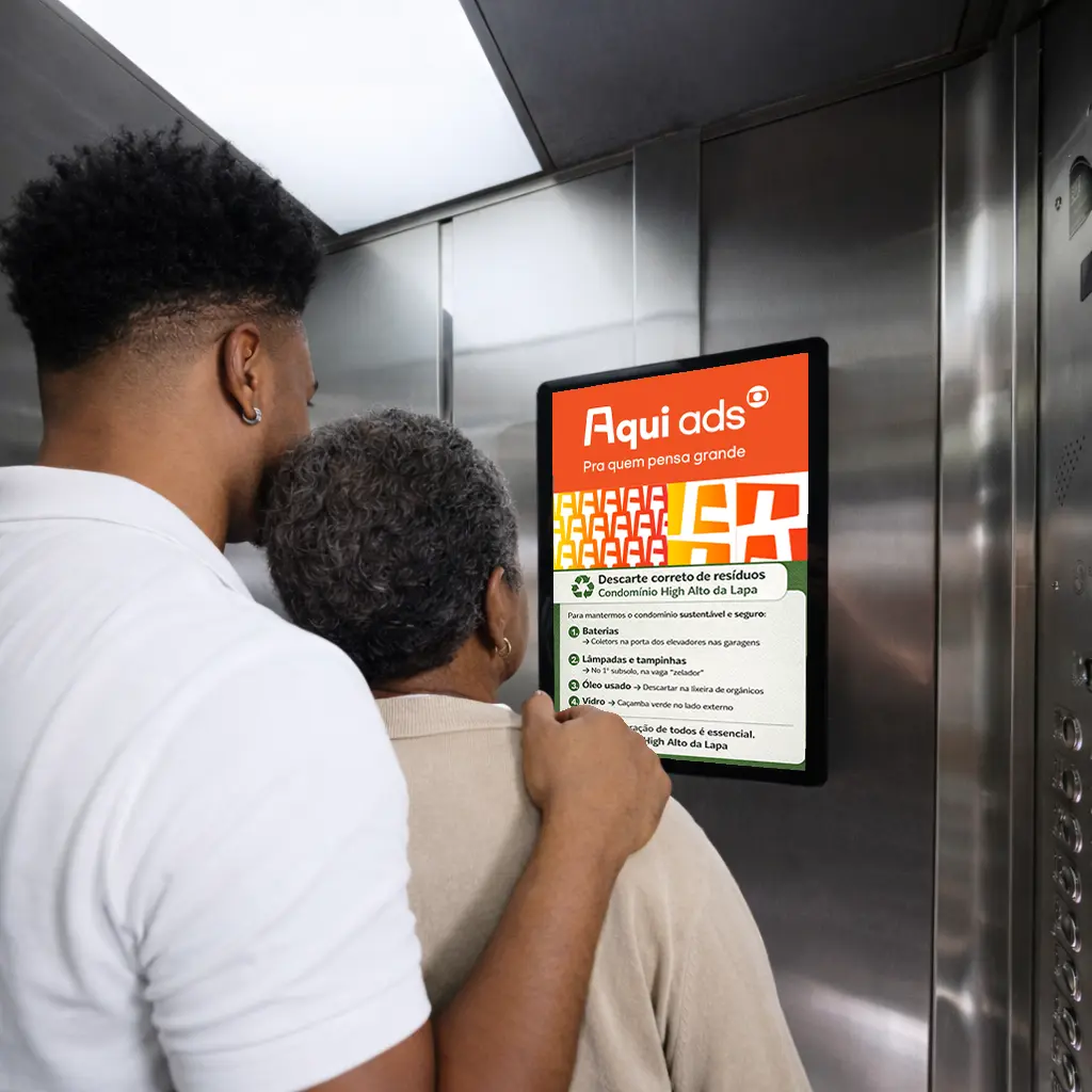 Um homem jovem e uma mulher mais velha, vistos de costas dentro de um elevador, observam uma tela digital fixada na parede de inox que exibe um anúncio da 'Aqui ads' e um comunicado sobre descarte correto de resíduos do condomínio.