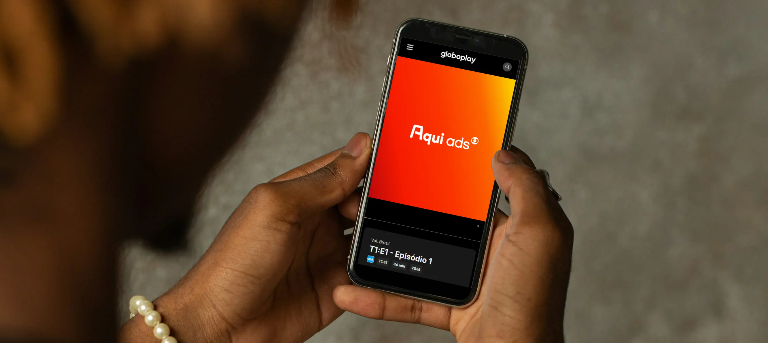 Mãos de uma pessoa segurando um smartphone que exibe a interface da plataforma Globoplay na tela. No centro do visor, um anúncio com fundo gradiente em vermelho e laranja destaca a marca 'Aqui ads'.