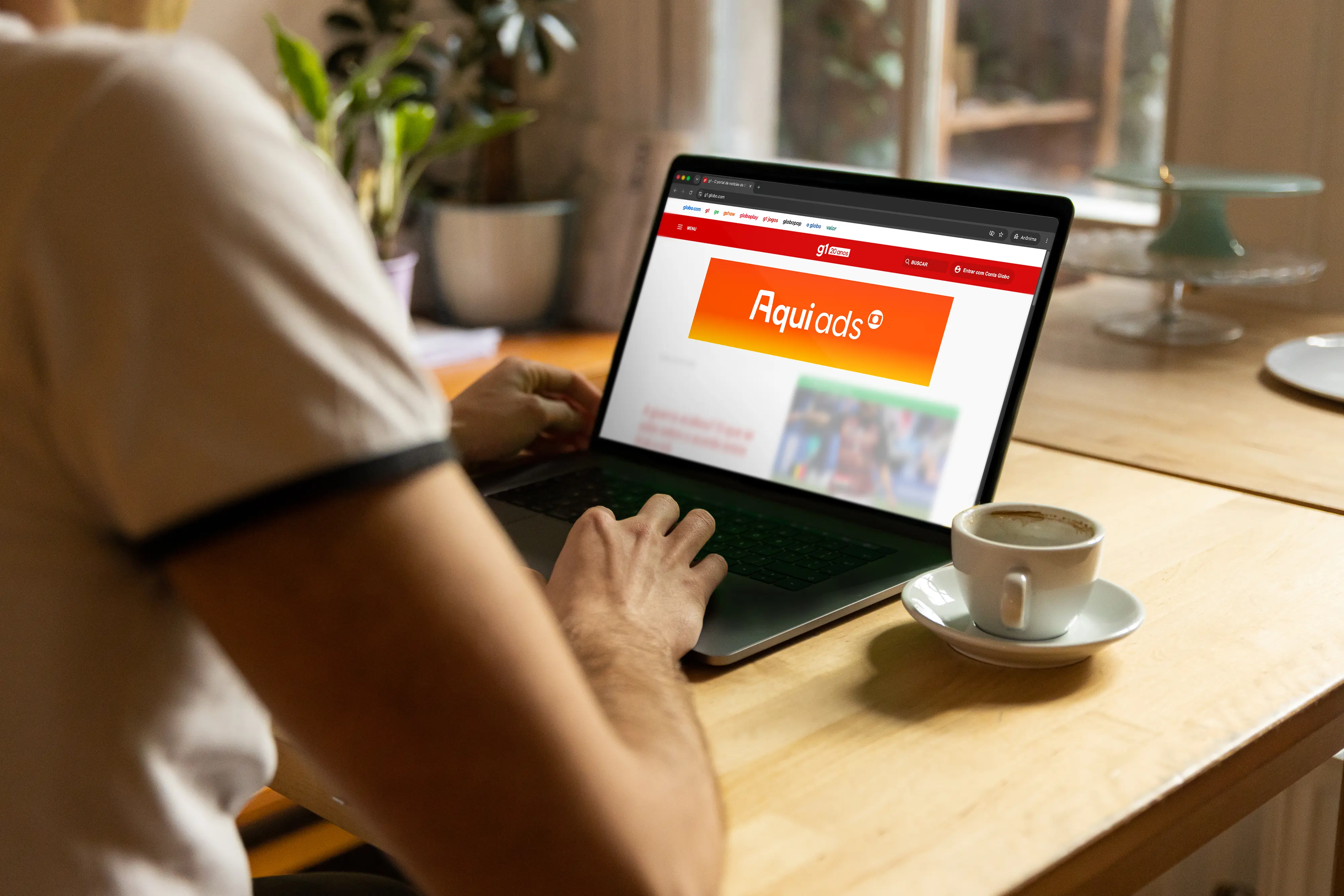 Uma pessoa sentada à mesa usa um notebook exibindo a página do portal g1. No topo do site, destaca-se um banner publicitário retangular em gradiente laranja com o texto 'Aqui ads'. Ao lado do computador, há uma xícara de café.